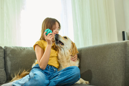 A girl with long hair enjoys her time at home with a golden retriever. She sits on a sofa, holding a green ball while the dog eagerly interacts with her, displaying a playful moment.の写真素材