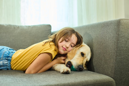 A young girl lies on a couch, playfully interacting with her golden retriever. The sunlight illuminates the room, creating a warm and inviting atmosphere.の写真素材