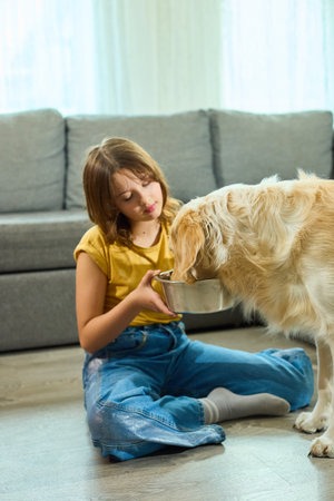 A teen girl sits on the floor in a welcoming living room, joyfully watching her golden retriever eat from a stainless steel bowl. The warm atmosphere invites companionship and happiness.の写真素材
