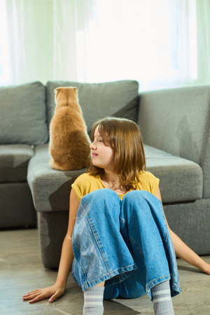 Girl sits on the floor while Scottish Fold cat relaxes on the couch in a cozy home environmentの写真素材
