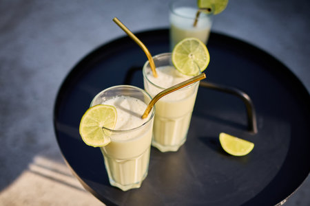 Glasses of lime and condensed milk cocktails sit on a black table in a sunlit terrace. Fresh lime wedges garnish each drink, highlighting a casual summer vibe.の写真素材