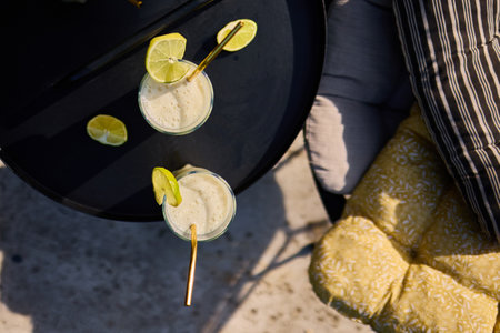 Glasses of lime and condensed milk cocktails sit on a black table in a sunlit terrace. Fresh lime wedges garnish each drink, highlighting a casual summer vibe.の写真素材