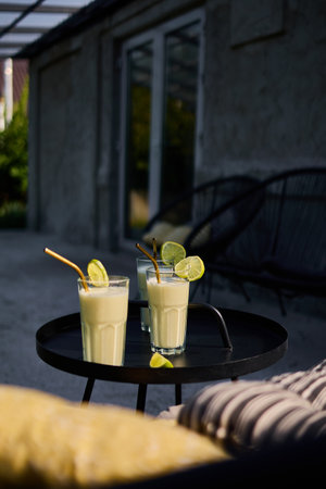 Glasses of lime and condensed milk cocktails sit on a black table in a sunlit terrace. Fresh lime wedges garnish each drink, highlighting a casual summer vibe.の写真素材