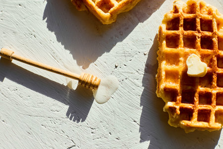 Waffles are arranged on a light surface, with one piece showing a small pat of butter and a honey dipper beside them, showing a tasty breakfast in natural morning light.の写真素材
