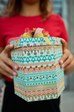 A child stands outdoors in front of a house, holding a colorful wrapped gift with a ribbon. Potted plants are visible in the background. It looks like a special occasion.の写真素材