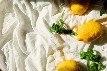 A collection of fresh lemons and vibrant basil leaves is artistically arranged on a light fabric...