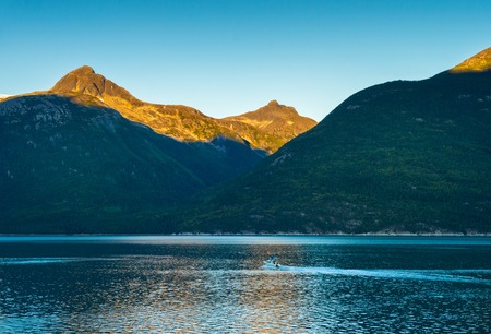 Small boat and sunrise light on mountains at dawn on calm early morning in beautiful Taiya Inlet,...