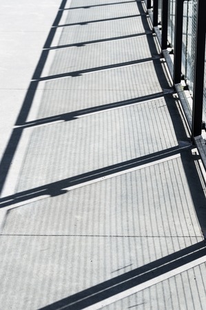 Diagonal shadows on cement walkway from metal and glass panel barrier.