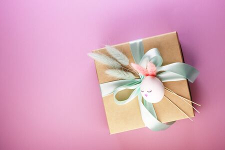 a gift box and a flower on the pink table on top. a box of paper with a blue bow. Easter egg in the form of a rabbit with ears and a cute face on the box. flat recumbent style.の写真素材