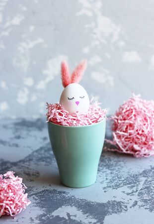 a white Easter egg with ears and a cute face in a pink nest. egg in the form of a rabbit in a nest of decorative paperの写真素材