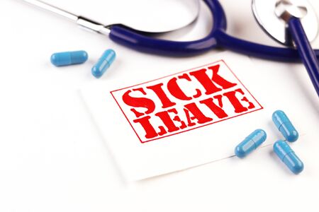 phonendoscope and a sheet with the text of the sick leave on a white background. medical blue capsules are scattered on the tableの写真素材