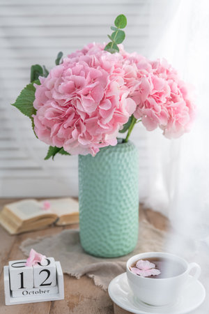 Composition with tea mug and beautiful pink flowers hydrangeas in vase, on wooden table, on white...