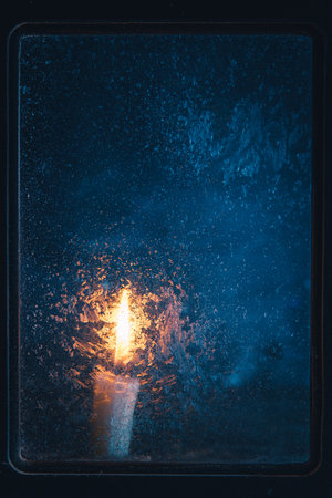 Christmas, new year, winter composition. Candle behind a frozen window with frosty patterns, copy spaceの写真素材
