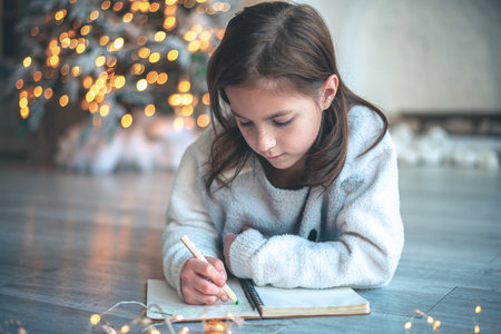 a cute girl lies on the floor near the Christmas tree and writes a letter to Santa Claus, a to-do list for the next yearの写真素材