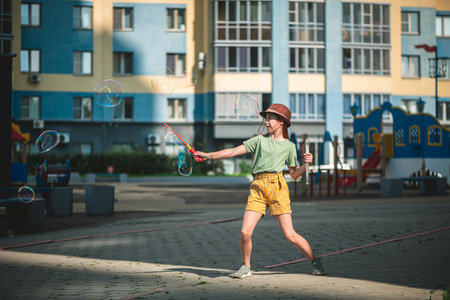 Cute school-age girl blows big soap bubbles in the courtyard of a multi-storey building in summer. summer concept, outdoor gamesの写真素材
