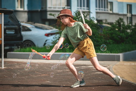 Cute school-age girl blows big soap bubbles in the courtyard of a multi-storey building in summer. summer concept, outdoor gamesの写真素材
