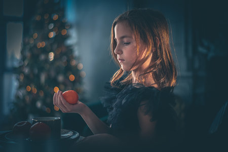 a cute girl in a black dress sits at a table with tangerine on the background of the Christmas tree. holiday, new year, christmasの写真素材
