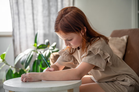 cute little preteen girl drawing with pen while sitting at desk at home, beautiful female child, small kid doing school homeworkの写真素材