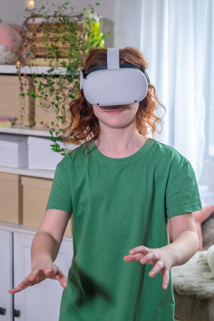 redhead teen girl at home in living room put on vr helmet to enjoy virtual reality online game cyber space world simulation. cute girl has fun using new electronic device for entertainmentの写真素材