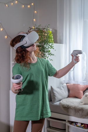 redhead teen girl at home in living room put on vr helmet to enjoy virtual reality online game cyber space world simulation. cute girl has fun using new electronic device for entertainmentの写真素材