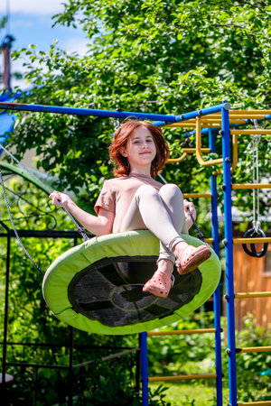Cute beautiful teenage girl with red hair swings on a swing in summer countryside, vacation, summertimeの写真素材