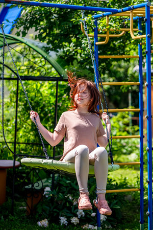 Cute beautiful teenage girl with red hair swings on a swing in summer countryside, vacation, summertimeの写真素材