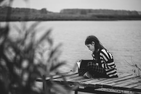 Pensive teen girl sitting on the pier by the river with a bookの写真素材