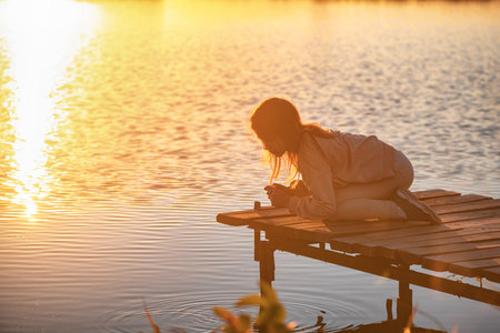 A cute girl teen sits on a pier, bridge by the river at sunsetの写真素材