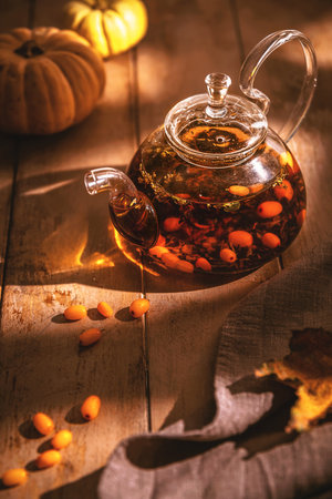 Glass teapot with sea buckthorn tea sits on a rustic wooden table, accompanied by pumpkins, scattered sea buckthorn berries, and autumn leaves, creating a cozy autumnal sceneの写真素材