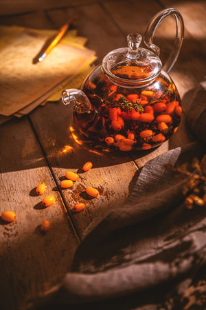 Glass teapot with sea buckthorn tea sits on a rustic wooden table, accompanied by pumpkins, scattered sea buckthorn berries, and autumn leaves, creating a cozy autumnal sceneの写真素材