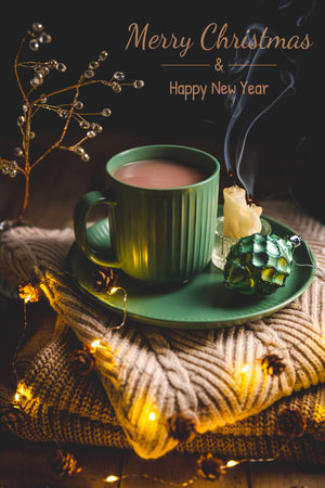 Steaming mug of hot chocolate resting on a green saucer beside an extinguished candle and ornament, placed on a cozy sweater adorned with warm fairy lights, creating a comforting winter sceneの写真素材