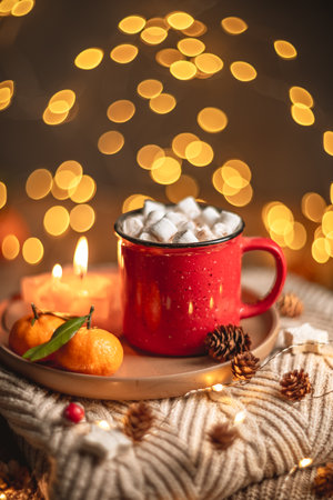 Red mug filled with hot chocolate and marshmallows resting on a tray with tangerines, surrounded by warm knit fabric and glowing Christmas lights, creating a cozy winter sceneの写真素材
