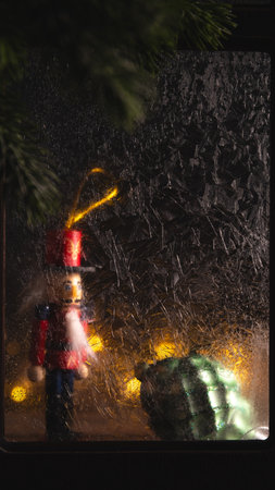 Wooden toy soldier and green christmas bauble are visible behind frozen window with warm fairy...