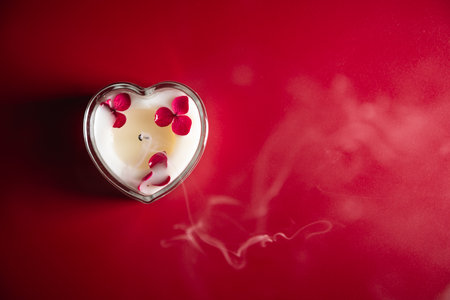Heart-shaped candle with red petals on a smooth red surface, emitting gentle smoke, creating a romantic and intimate atmosphere perfect for Valentine's day celebrationsの写真素材