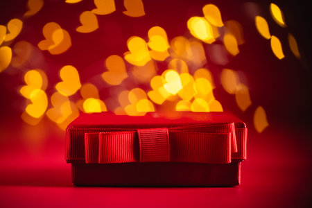 Red gift box adorned with a ribbon, set against a backdrop of glowing heart-shaped bokeh lights, creating a romantic atmosphere ideal for Valentine's day celebrationsの写真素材