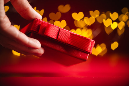 Hand gracefully presenting a red gift box adorned with a ribbon, set against a backdrop of glowing, heart-shaped bokeh lights, symbolizing love and celebration on Valentine's dayの写真素材