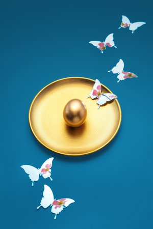 A gold egg on gold plate with mirror butterflies, which reflects pink spring flowers, festive atmosphere of Easter spring on a bright turquoise background. Easter creative luxury postcardの写真素材