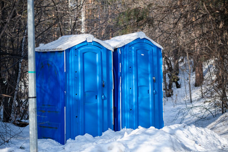 Two blue portable toilet cabins are standing on a snowy ground in winter, surrounded by leafless trees and bushes, providing necessary facilities in outdoor environmentsの写真素材