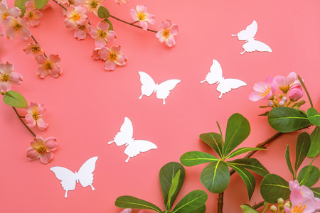 Delicate white paper butterflies are artfully arranged among pink blossoms and lush green leaves on a vibrant coral background, creating a whimsical and enchanting spring or easter sceneの写真素材