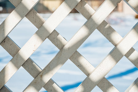 Sunlight streaming through a white wooden lattice creates diamond-shaped openings, revealing a blurred, snow-covered background in a serene winter settingの写真素材