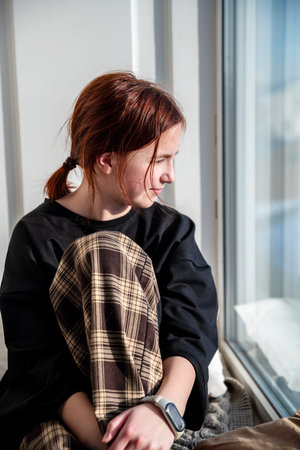 Redhead teenage girl with plaid trousers sitting next to window enjoying sunlight and contemplating life, feeling peaceful and sereneの写真素材
