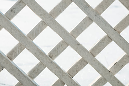 Sunlight streaming through a white wooden lattice creates diamond-shaped openings, revealing a blurred, snow-covered background in a serene winter settingの写真素材