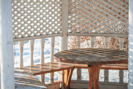 Sunlight streaming through the lattice of a white gazebo, illuminating a wooden table and benches while casting intricate shadows on the snow-covered floor during winterの写真素材