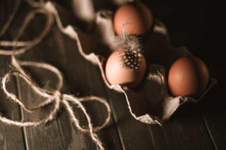Fresh brown eggs sit in a cardboard carton on a rustic wooden table, one adorned with a delicate guinea fowl feather, creating a simple yet elegant composition, top viewの写真素材