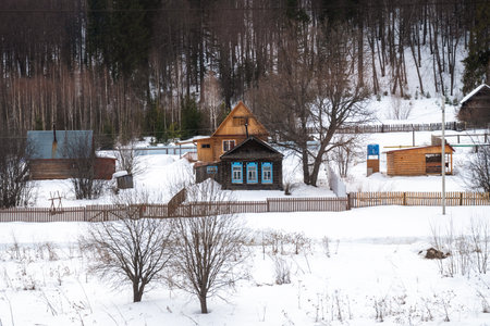 Snow covers the ground around traditional Russian wooden houses with blue painted window frames and wooden fences in a serene winter landscape, showcasing the rustic charm of rural lifeの写真素材