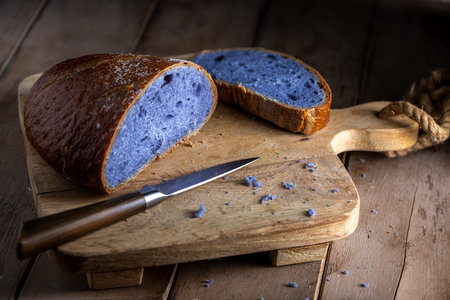 Freshly baked purple bread made with Thai blue ancha tea, partially sliced and resting on a rustic wooden cutting board, looks attractive next to a knife, ready to serve and eat.の写真素材