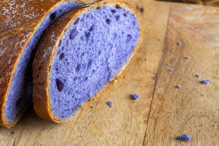 Freshly baked bread with a vibrant purple interior, cooked in blue Thai ancha tea, rests on a rustic wooden cutting board with a slice cut off to reveal its soft, crumbly texture, with copy spaceの写真素材