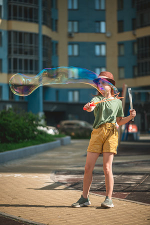 Cute school-age girl blows big soap bubbles in the courtyard of a multi-storey building in summer....