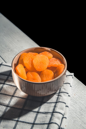 Delicious dried apricots arranged in a bowl resting on a checkered cloth atop a white wooden table, offering a nutritious snack or a flavorful ingredient for cookingの写真素材