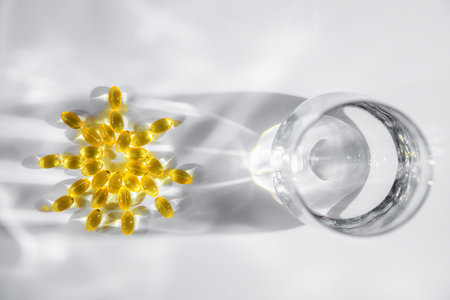 vitamin D or Fish oil yellow transparent capsules forming a sun shape alongside a glass of water create playful shadows on a white background, emphasizing themes of health, wellness, and nutritionの写真素材